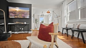 Un salon moderne avec un grand téléviseur à écran plat, un fauteuil en osier avec un coussin rouge, une table basse en bois et un tapis blanc.