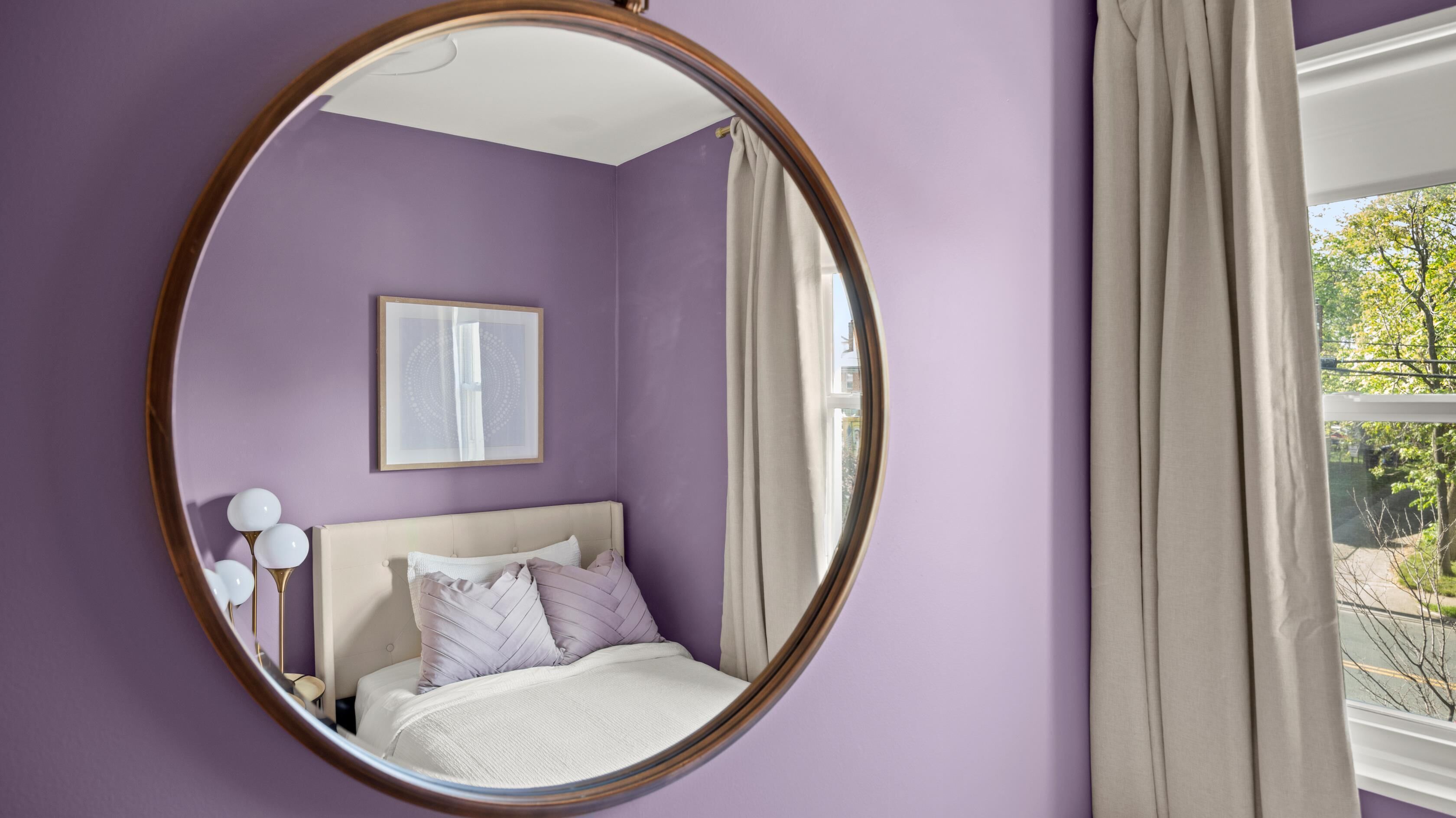 Une chambre avec un miroir rond, un lit avec des oreillers, un tableau encadré au mur et une fenêtre avec des rideaux.