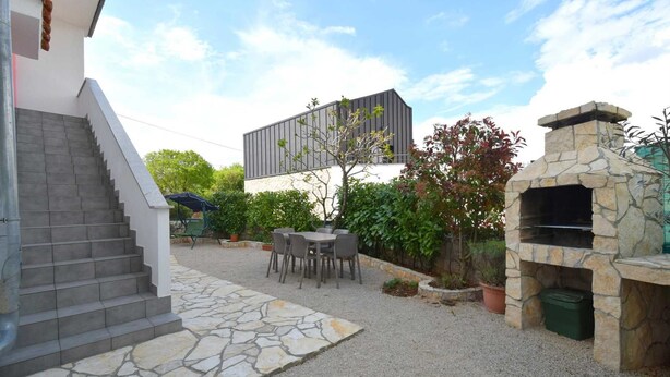 A patio area with a stone staircase, a table with chairs, a stone barbecue grill, and a modern building in the background. - Studio-apartment Letizia (Rasopasno)