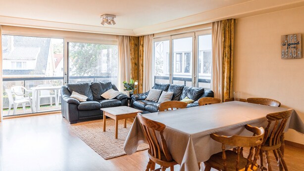 Ett modernt vardagsrum med en soffa, ett matbord och stolar. Stora fönster ger naturligt ljus och en utsikt över omgivningen.