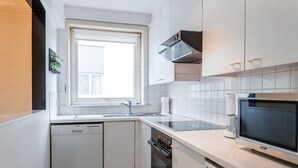 Ett kompakt kök med vita skåp, en inbyggd ugn, en diskmaskin och ett fönster som släpper in naturligt ljus.