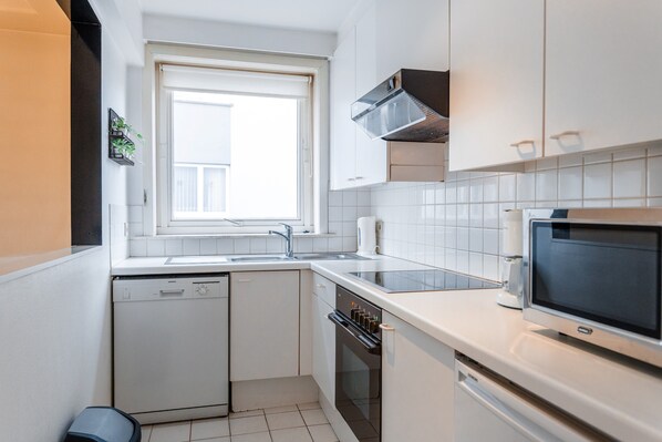 Una cocina compacta con armarios blancos, horno empotrado, lavavajillas y ventana que proporciona luz natural.