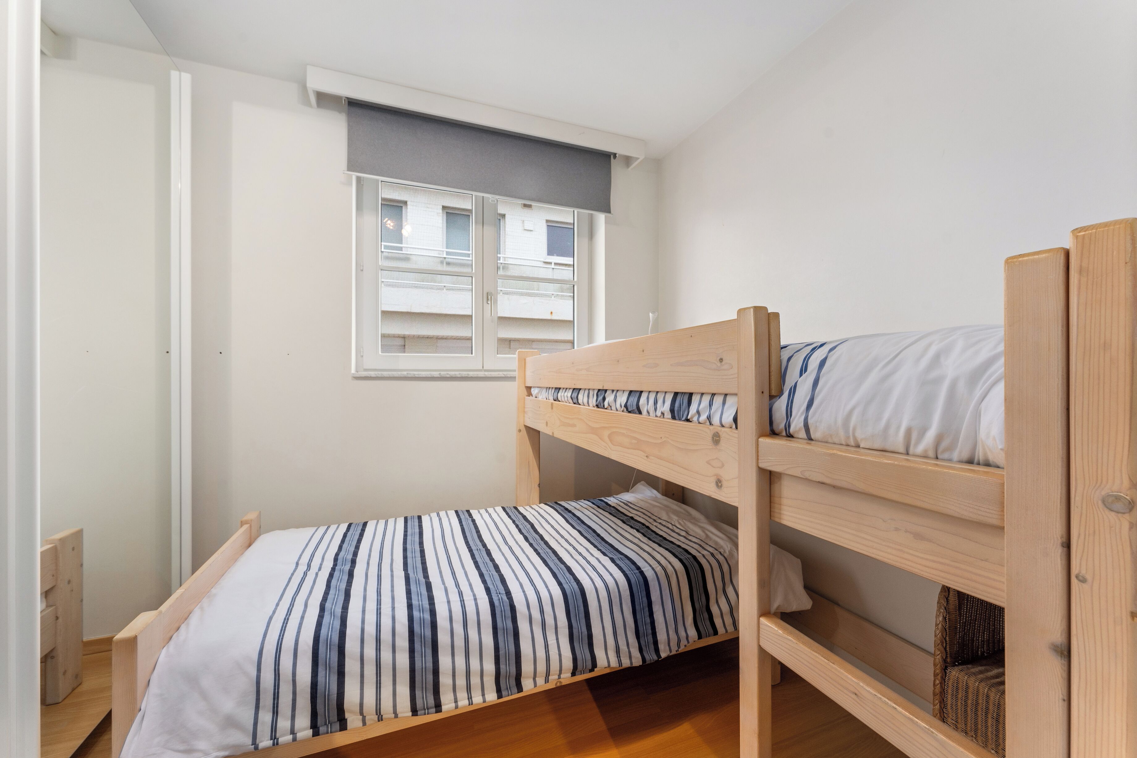 A small, single-room apartment with a bunk bed, a window with blinds, and a small wooden cabinet.