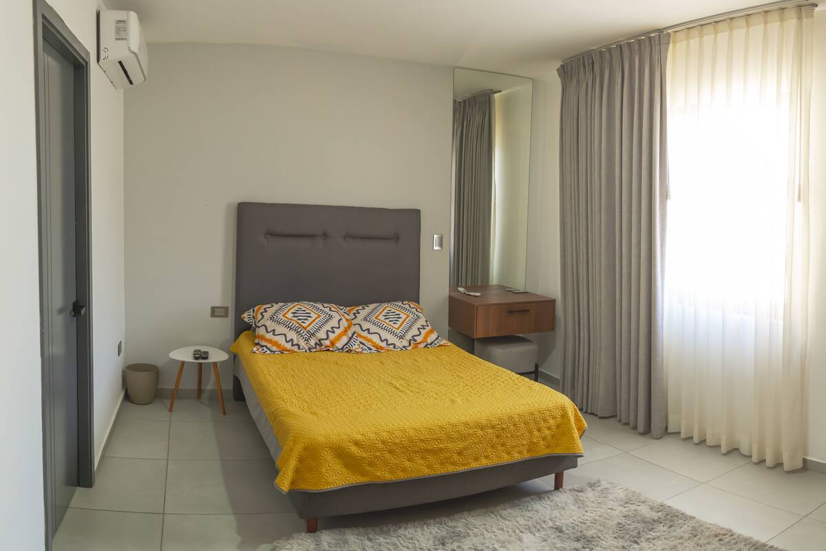 A bedroom with a bed, a bedside table, a mirror, and a window with curtains.