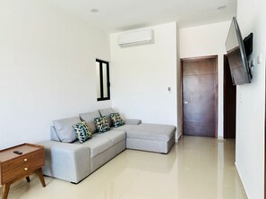 A modern living room with a grey sectional sofa, a wooden nightstand, and a flat-screen TV mounted on the wall.