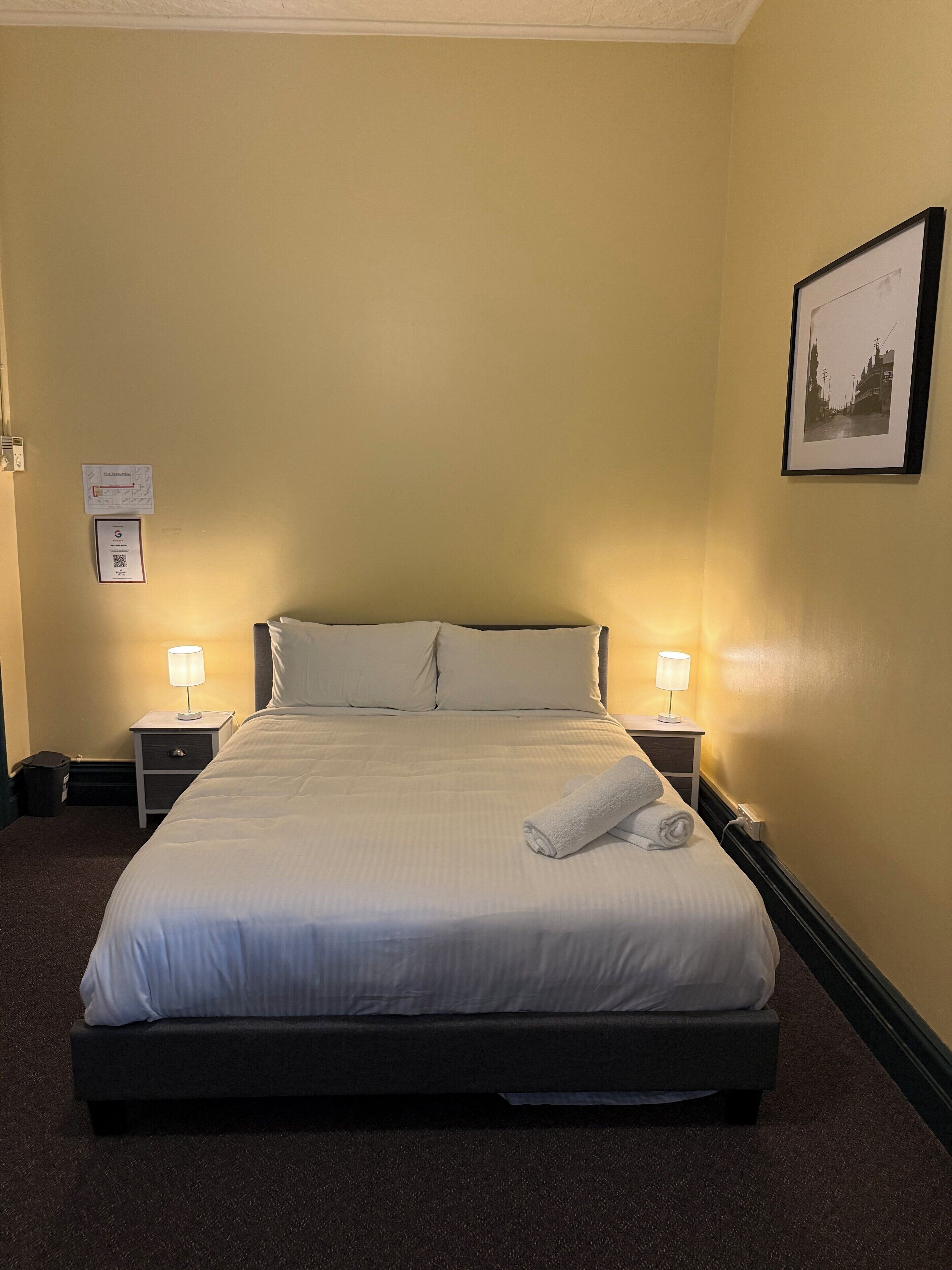 A neatly made bed with white linens, two bedside tables with lamps, a framed picture on the wall, and a small trash bin.