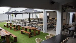 Een terras met houten tafels en banken, omringd door witte stoelen en beschut onder parasols.