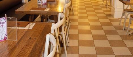 A dining area with a long wooden table, white chairs, and a checkered floor. There is a kitchen visible in the background.