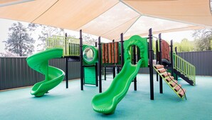 Een speeltuin met twee groene glijbanen, een klimstructuur en een vrolijke boomstam-brug.