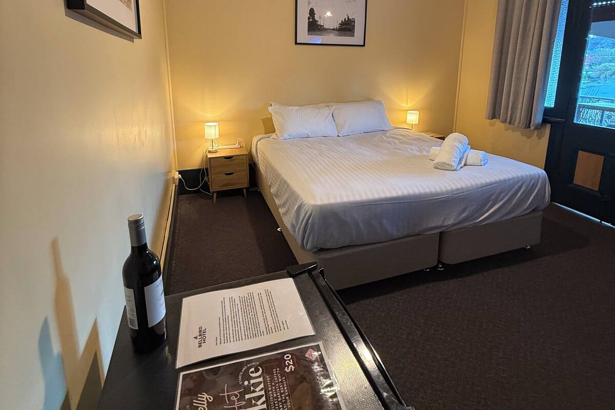 Habitación de hotel con cama, lámparas de noche, un cuadro enmarcado, ventana con cortinas y un menú sobre la mesa.