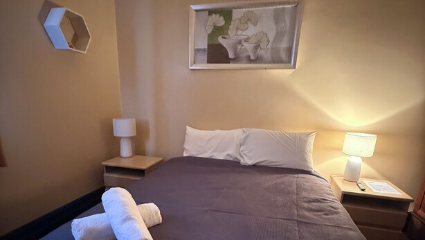Un lit bien fait, avec du linge de lit blanc et des oreillers, une table de chevet avec une lampe et une œuvre d’art encadrée au mur.
