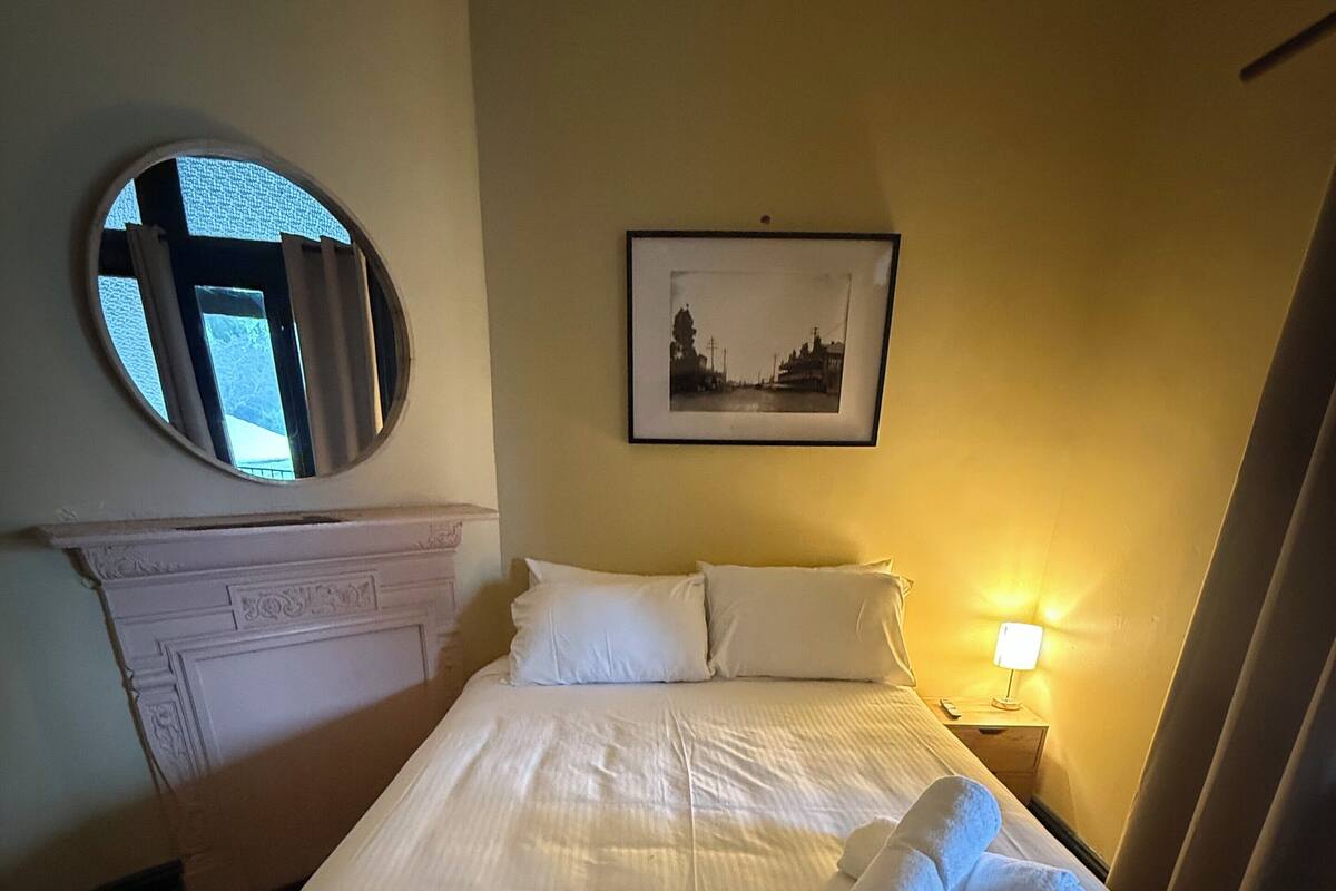 Una cama bien hecha con sábanas blancas y dos toallas, un cuadro en la pared, un espejo redondo sobre la cama y una mesita de noche con una lámpara.