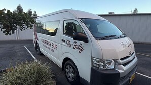 Een witte bestelwagen staat geparkeerd op een parkeerplaats met de aanduiding 'COURTESY BUS' en branding 'The Belly', omringd door planten en een heldere lucht.