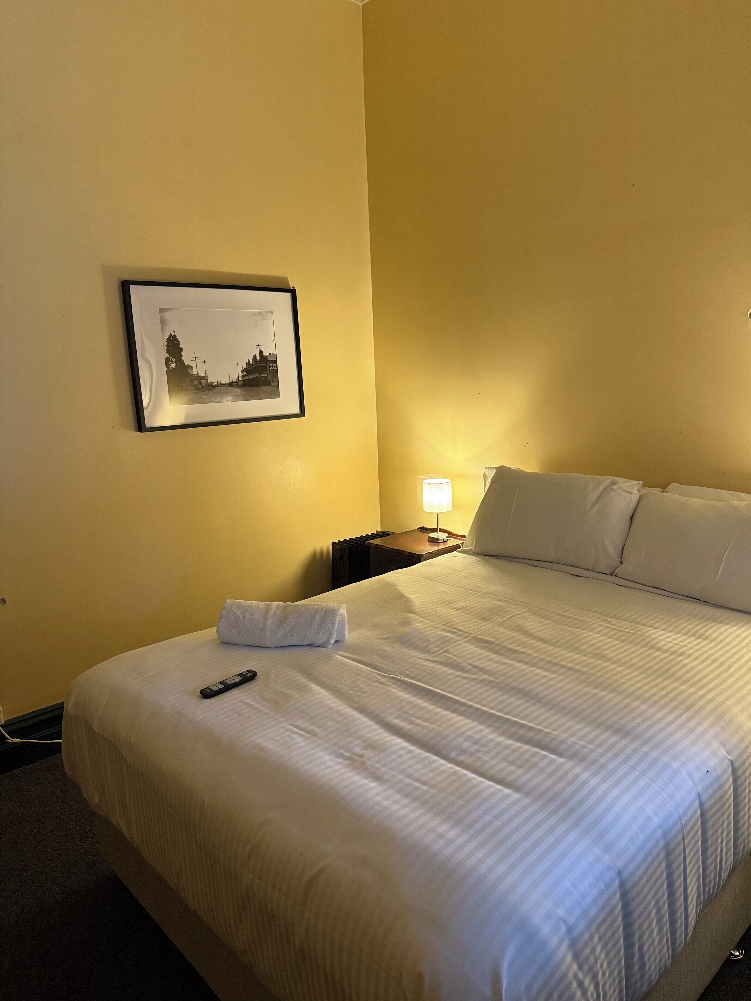 Ein Hotelzimmer mit einem ordentlich bezogenen Bett, einer Fernbedienung, einem gerahmten Bild an der Wand und einer Nachttischlampe.