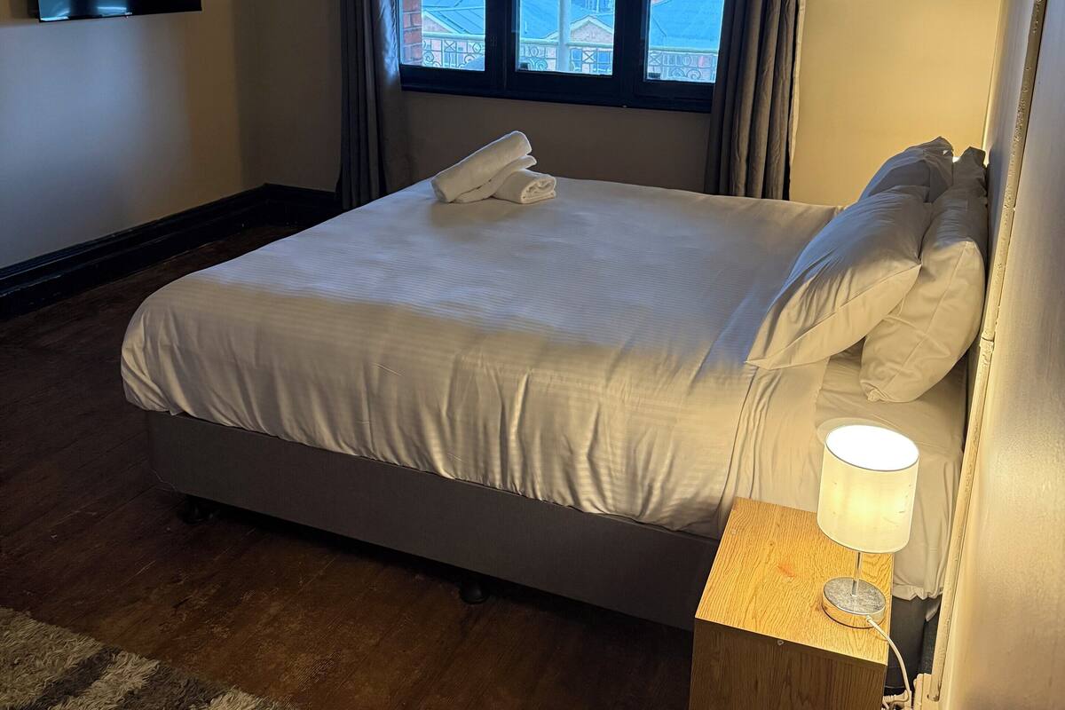 Habitación de hotel con cama, televisor montado en la pared, aire acondicionado, ventana con cortinas y lámpara sobre una mesita de noche.