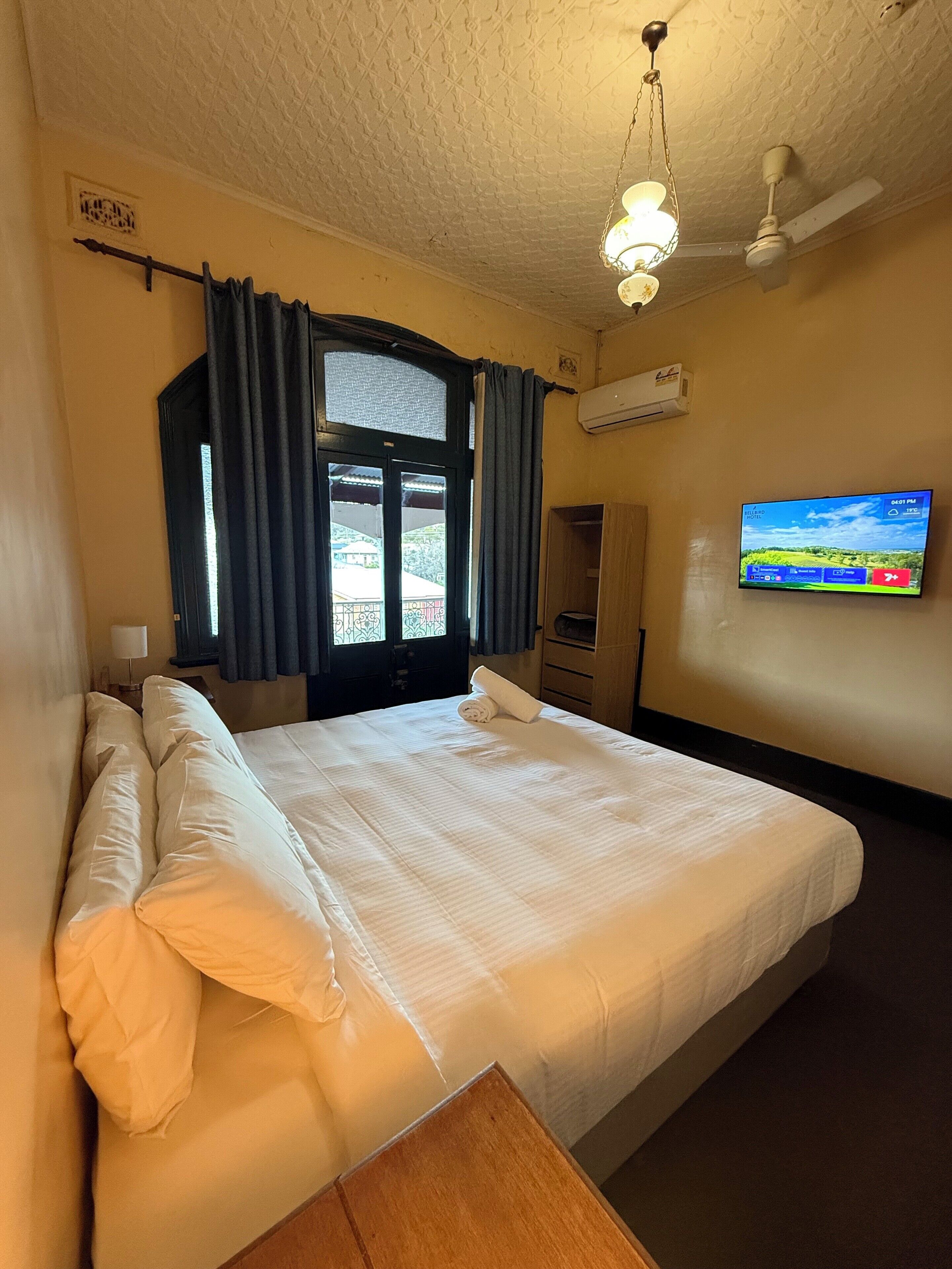 A hotel room with a bed, a TV mounted on the wall, a ceiling fan, and an air conditioning unit.