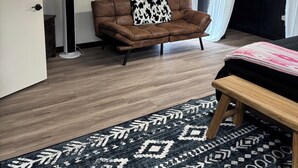 A room with a brown leather sofa, a black and white patterned rug, a wooden bench, a standing fan, and a ceiling light. - Perfect place to stay for events at the Lazy E (Guthrie)