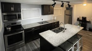A modern kitchen with dark cabinetry, stainless steel appliances, a granite countertop, and a bar stool seating area. - Current Bungalow 5 Beds Pro Turnkey MP Getaways (Clarksville)