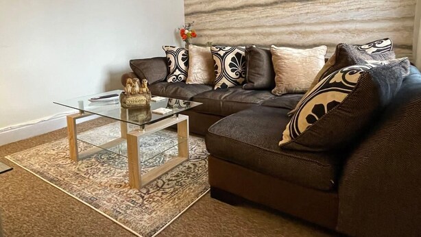 A living room with a brown sofa, patterned cushions, a glass coffee table, and a decorative wall piece. - Impeccable 4-Bed Home Away From Home in Swindon (Swindon)