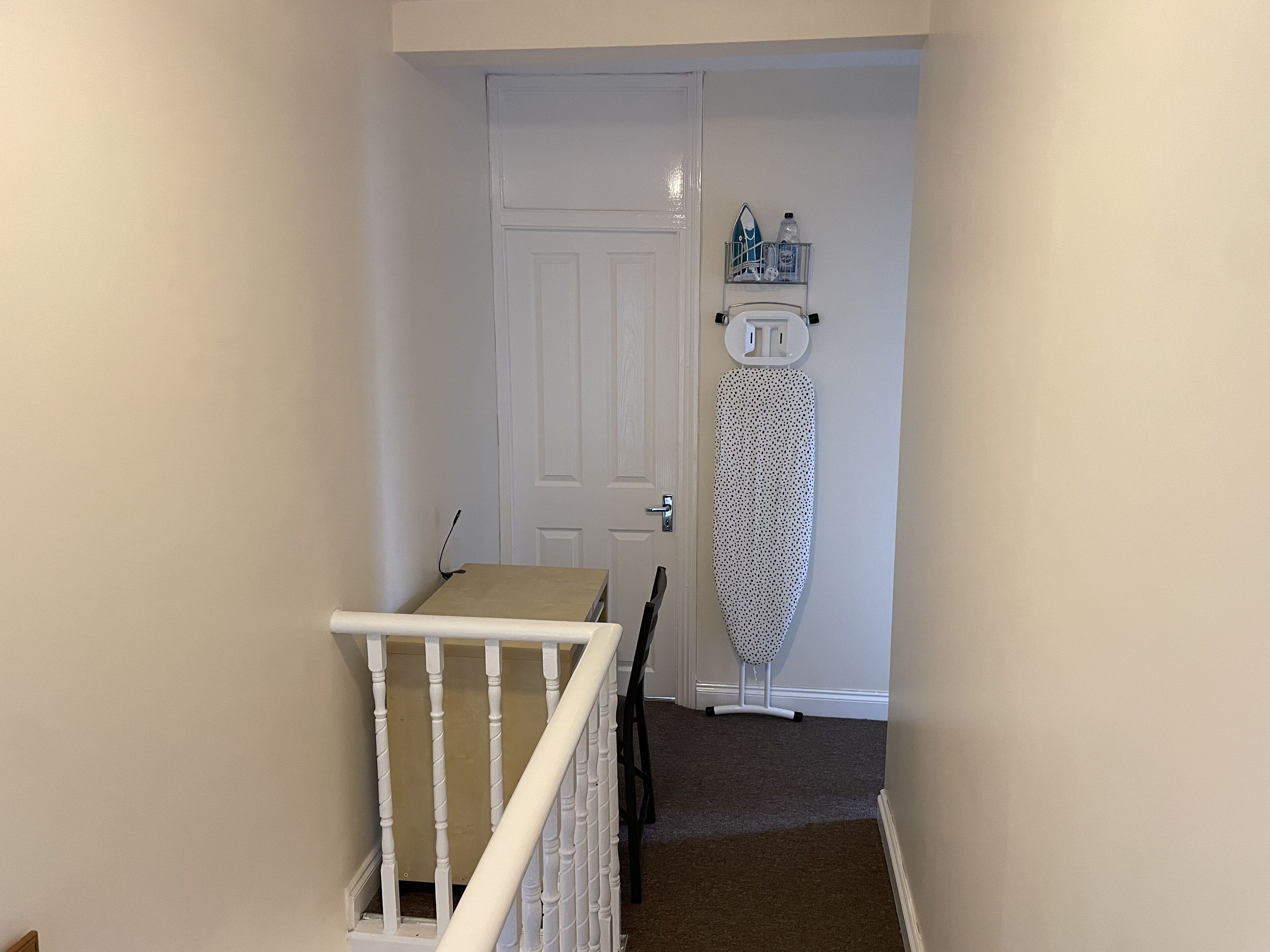 A narrow hallway with a white door, a small table with a laptop, and an ironing board against the wall.