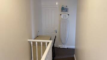 A narrow hallway with a white door, a small table with a laptop, and an ironing board against the wall.