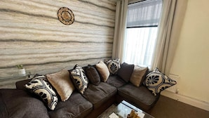 A living room with a brown leather sectional sofa, patterned throw pillows, a marble wall, a window with blinds, and a glass coffee table. - Impeccable 4-Bed Home Away From Home in Swindon (Swindon)