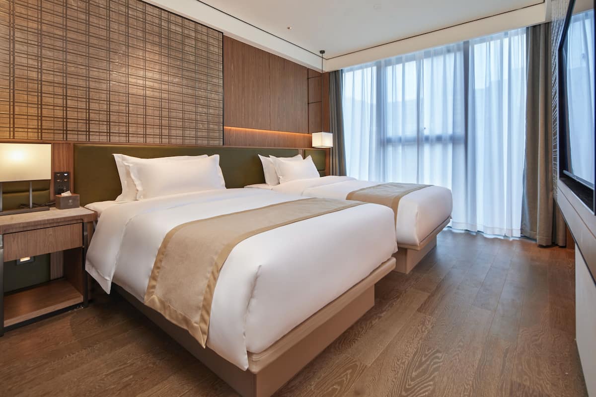 Ein Hotelzimmer mit zwei Betten, Holzboden, einem Nachttisch mit Lampe und einem großen Fenster mit Vorhängen.