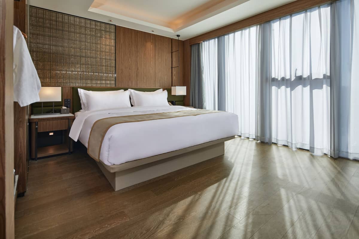Ein modernes Hotelzimmer mit einem großen Bett, Holzvertäfelung, einem Nachttisch und einem Fenster mit durchsichtigen Vorhängen.