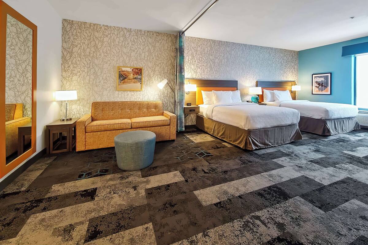 Hotellihuone, jossa on sohva, kaksi sänkyä, pyöreä jalkarahi, peili ja kuviollinen matto.
