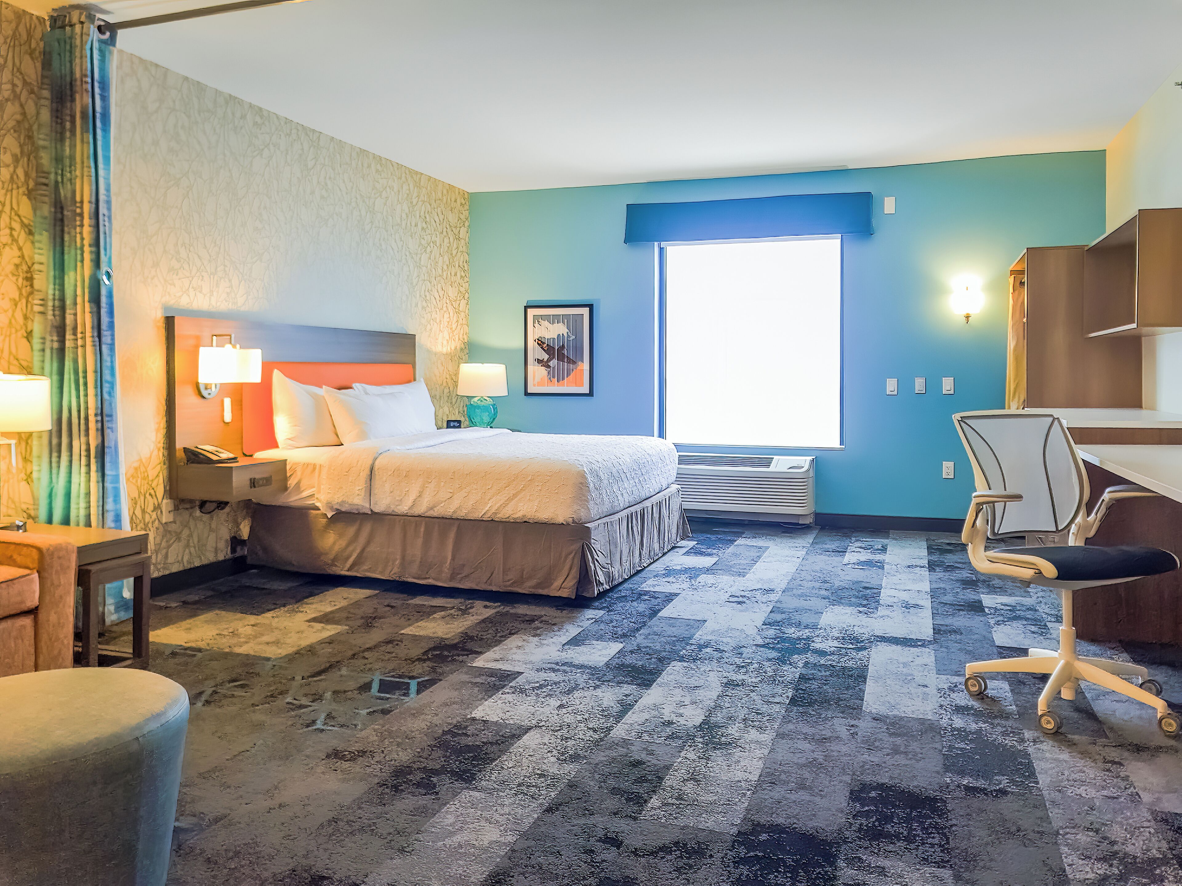 A hotel room with a bed, bedside lamps, a desk, a chair, and a large window.