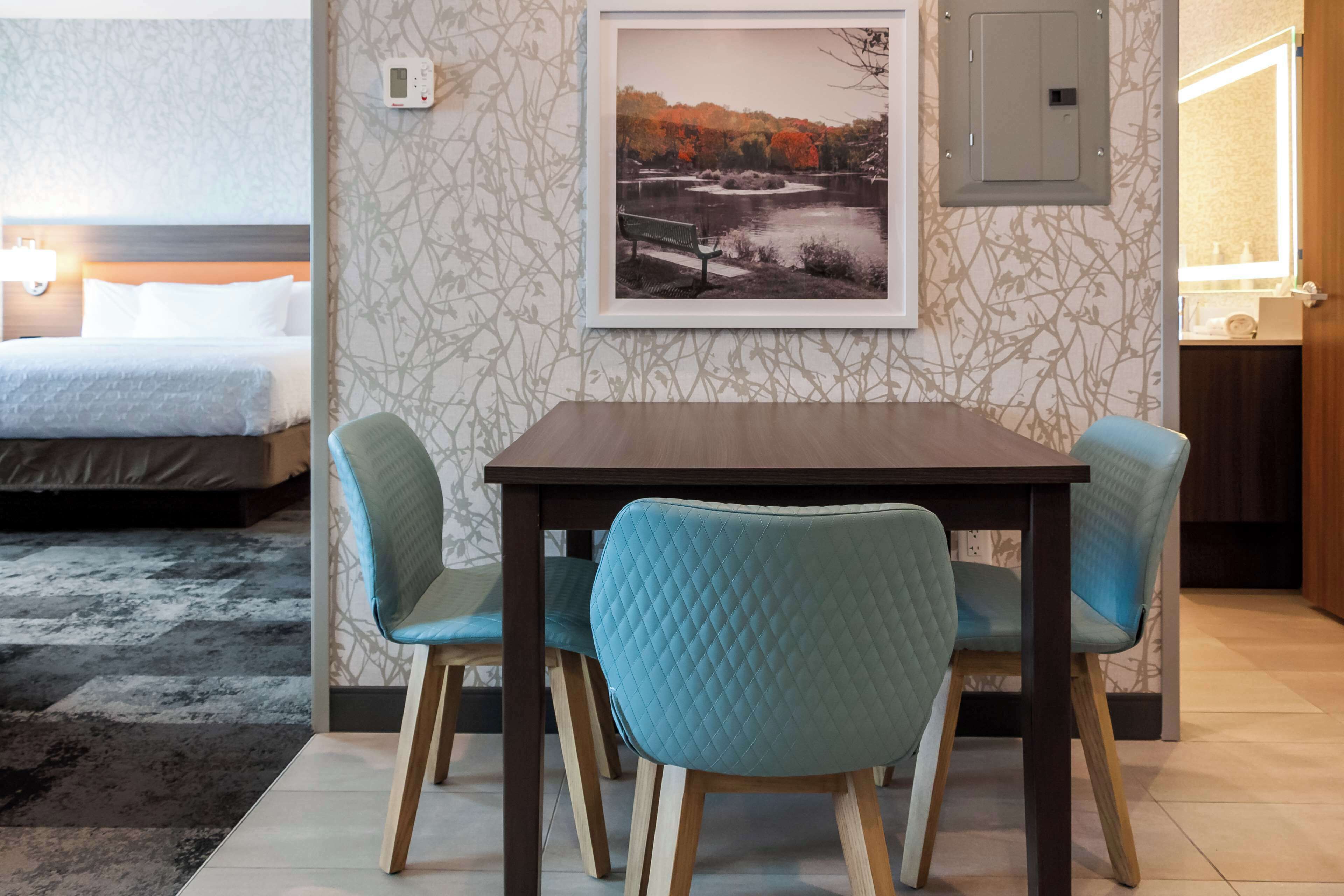 Hotellihuone, jossa on sänky, ruokapöytä tuoleineen, seinällä oleva kehystetty kuva ja avoimesta ovesta näkyvä kylpyhuone.