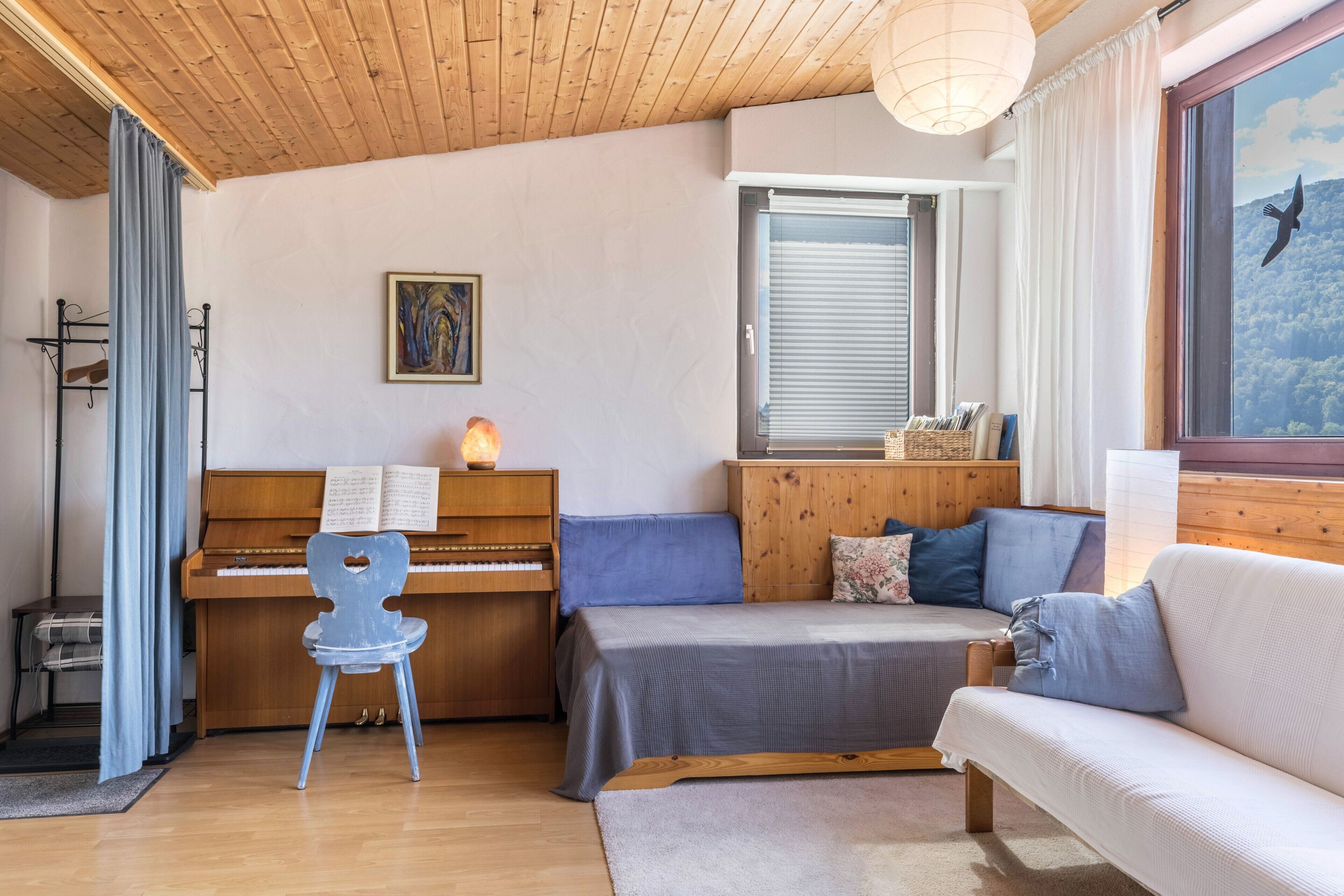 A cozy room with a piano, a bed, a sofa, and a window with a view of a forest.