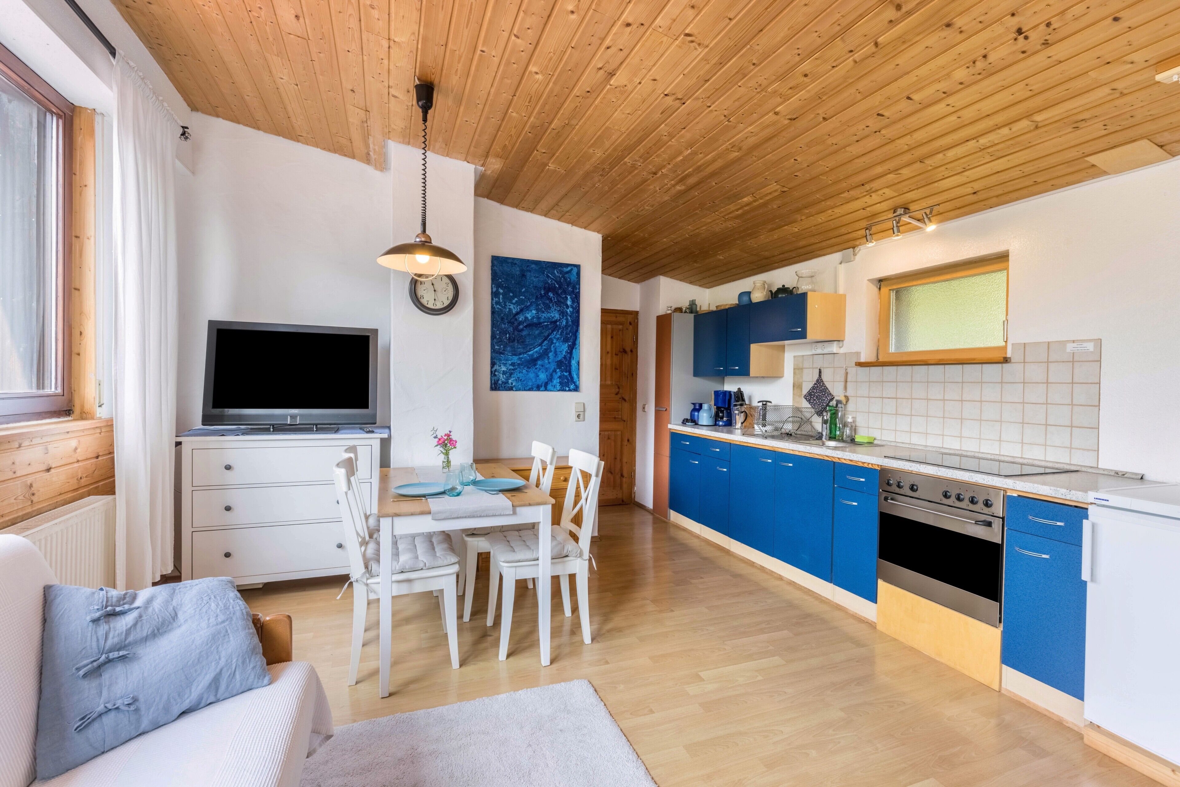 Ett modernt kök med blå skåp, en matplats med vita stolar, en tv och ett träpanelat tak.