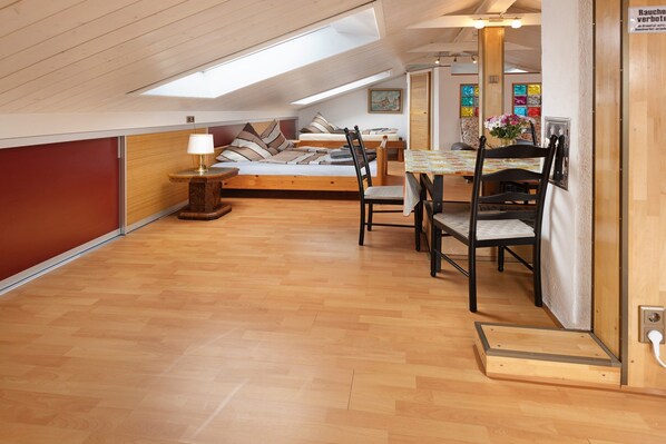 Un dormitorio estilo loft con tragaluces, suelo de madera, una cama con almohadas, un escritorio con silla y una mesa de comedor con sillas.