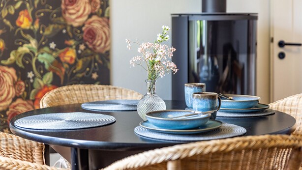 Ein Esstisch mit blauen Tellern und einer Blumenvase, umgeben von Korbstühlen.
