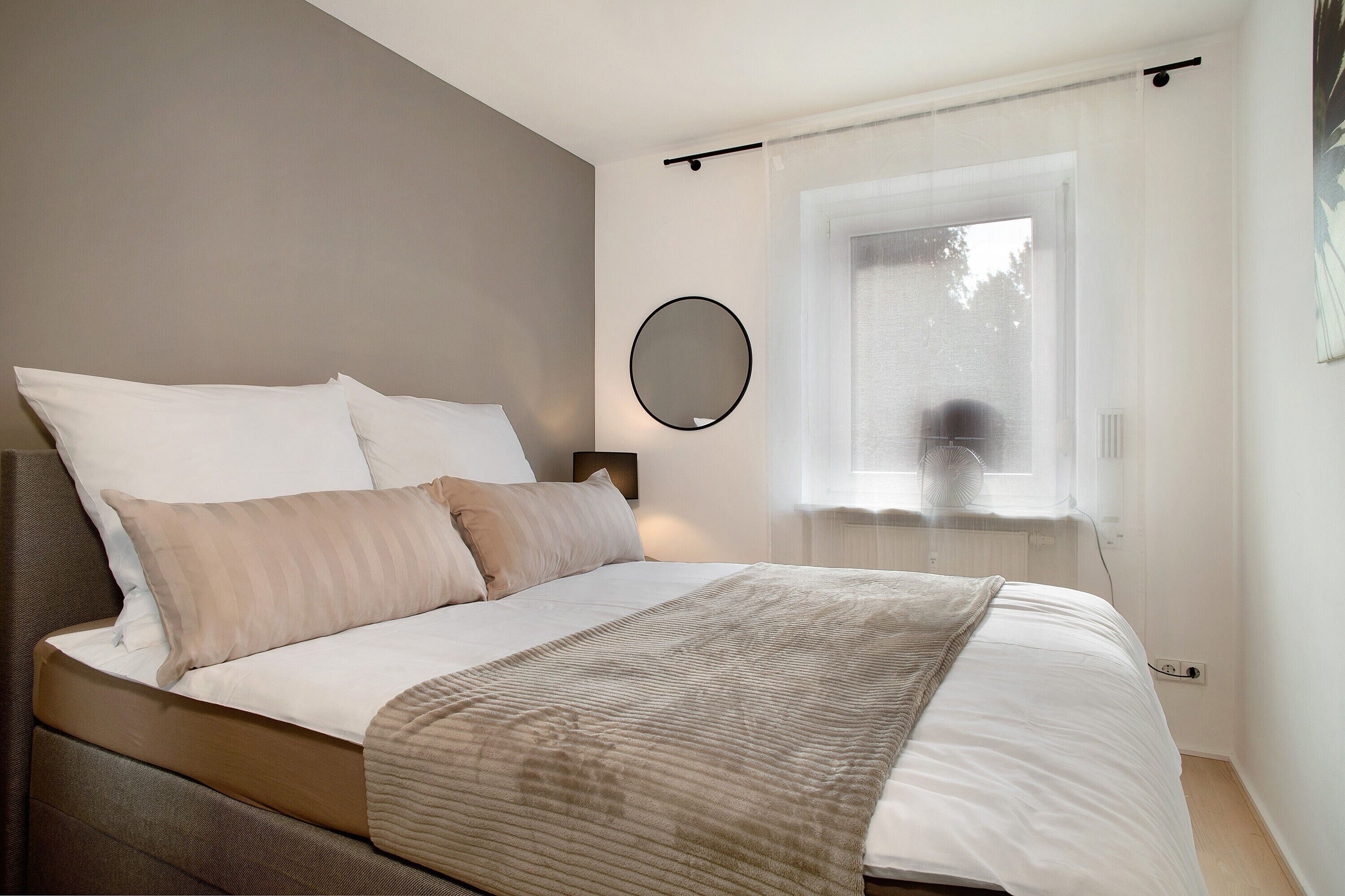 A neatly made bed with a headboard, a bedside table with a lamp, a round mirror on the wall, and a window with a view of trees.