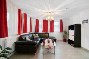 Un salón con un sofá de cuero negro, una mesa de centro de vidrio con camino de mesa floral, un mueble negro con cajones y cortinas rojas en las ventanas.