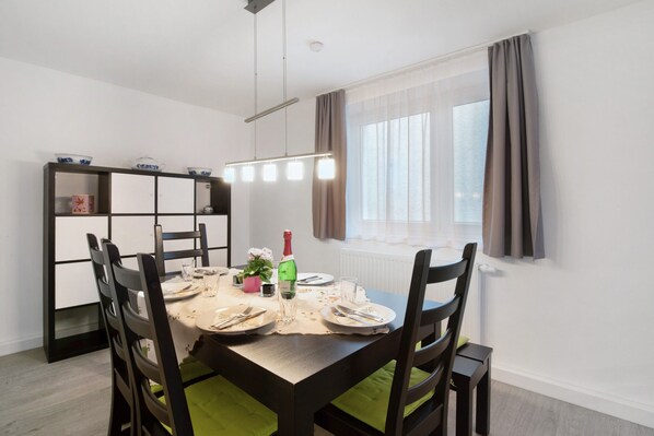 Un comedor con una mesa dispuesta para cuatro, una estantería y una ventana con cortinas.