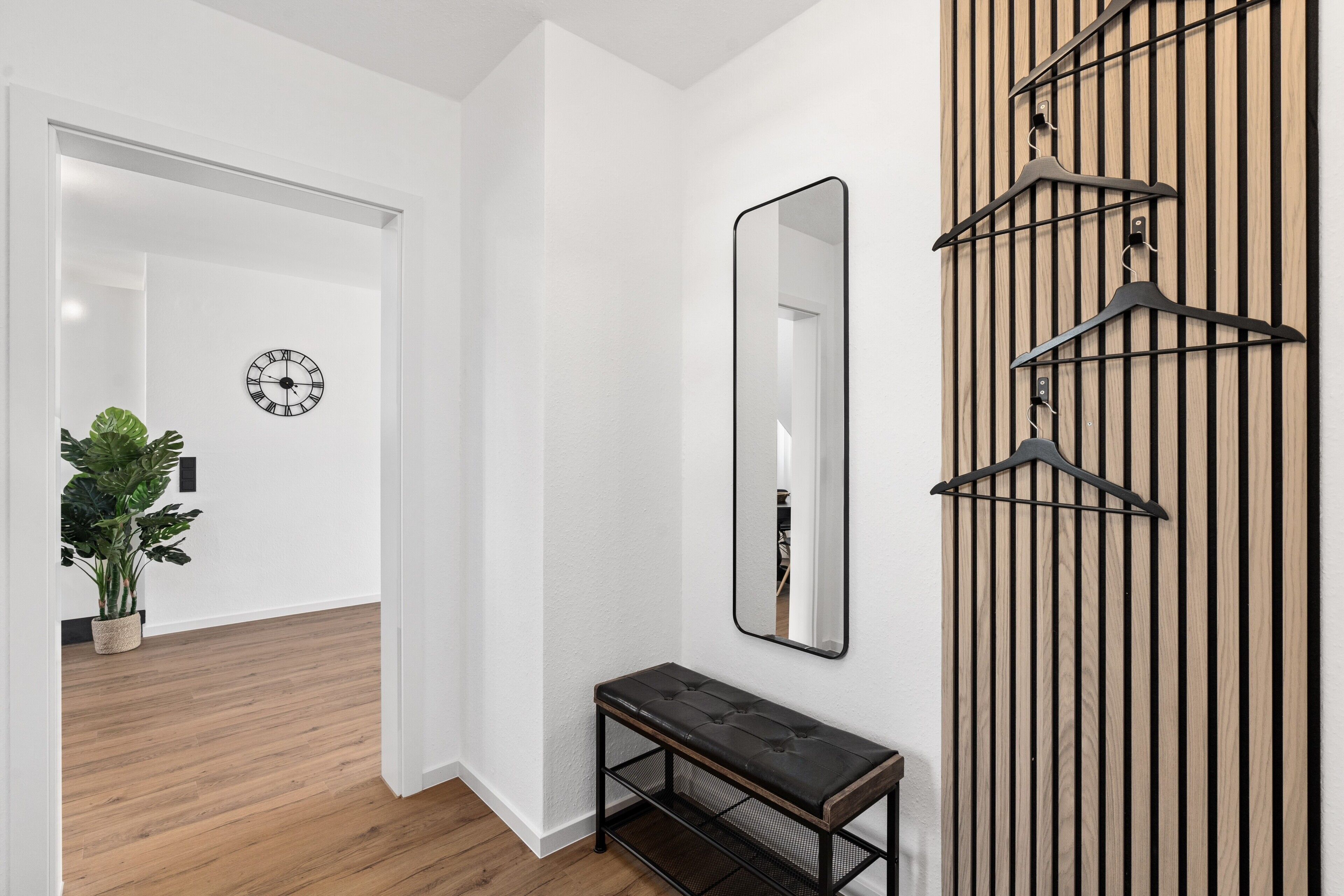 Un pasillo con suelo de madera, un banco, un espejo, un reloj y un panel de madera con objetos colgando.