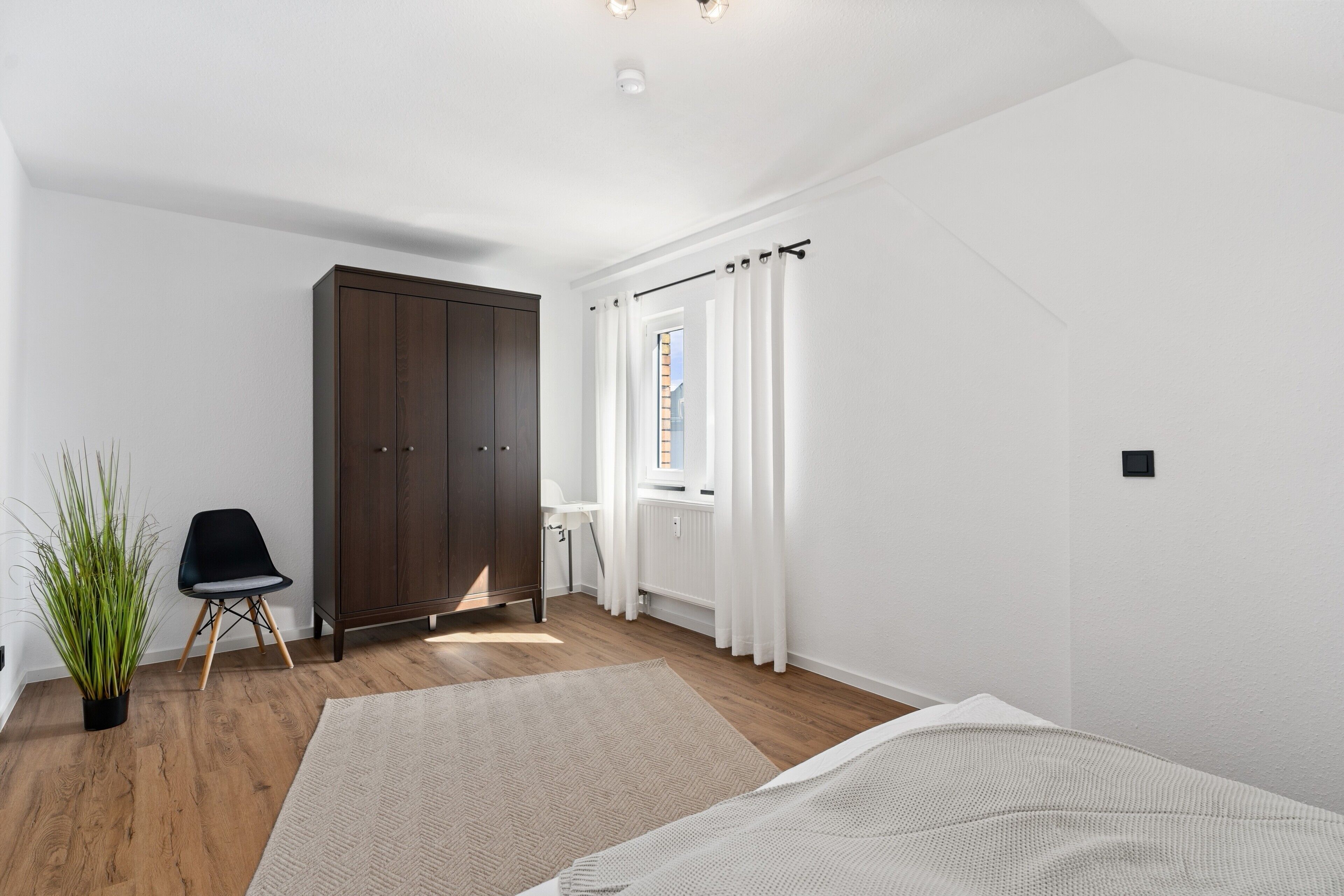 A room with a wooden floor, a large wardrobe, a chair, a potted plant, and a window with curtains.