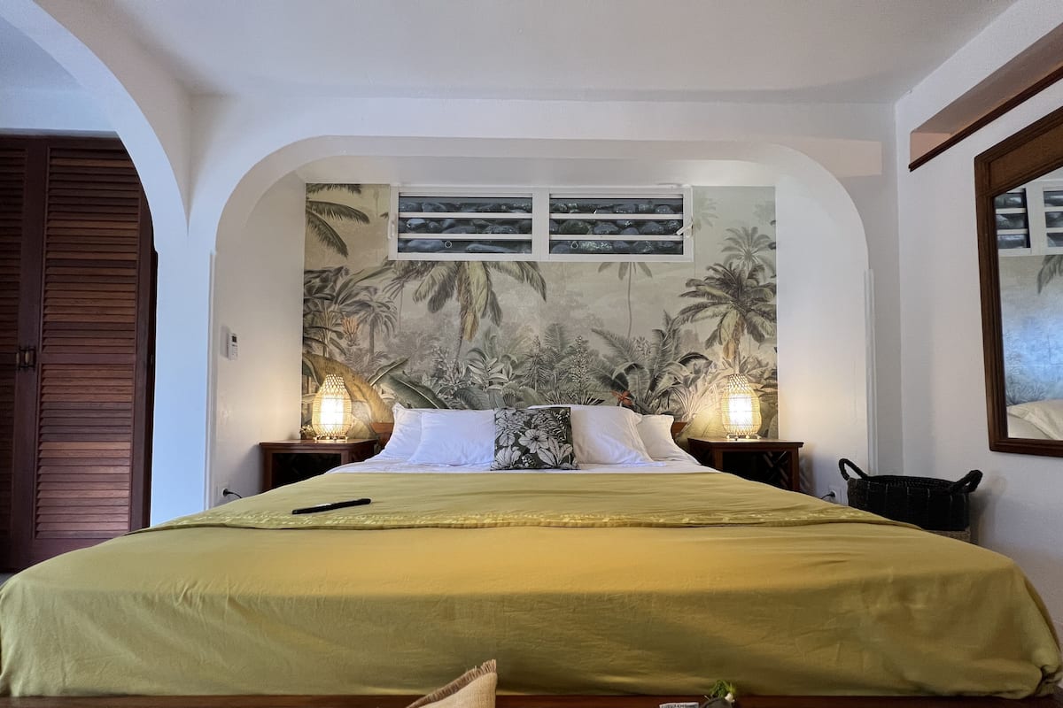 A large bed with a green bedspread, two bedside lamps, and a tropical mural on the wall.