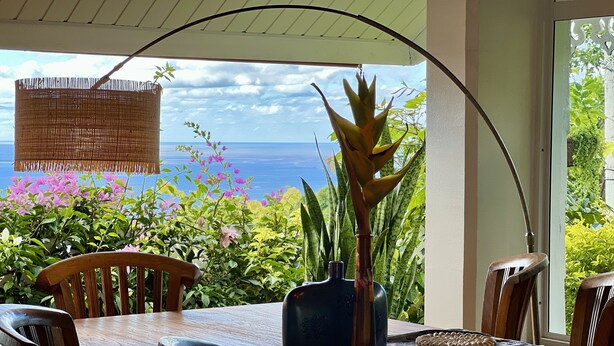 Une table à manger en bois, des chaises, un vase avec une plante et une vue sur l’océan.
