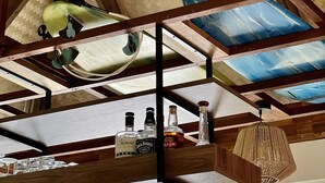 Une pièce avec un plafond haut, des poutres en bois et un plafond en osier tressé. On y trouve des étagères remplies de bouteilles et de verres, un luminaire suspendu et une fenêtre offrant une vue agréable.