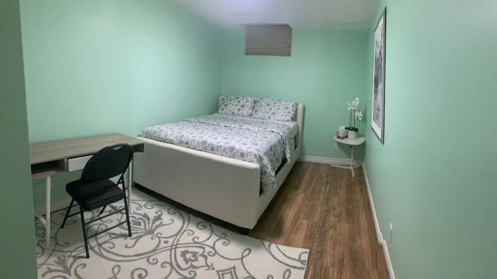 A bedroom with a bed, a desk, a chair, a small table with a plant, and a framed picture on the wall.