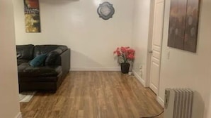A hallway with wooden flooring, a black sofa with cushions, a wall-mounted decorative plate, a potted plant with red flowers, and a radiator.