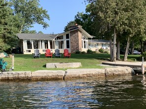 Ett hus vid sjön med en fasad av sten, en täckt veranda med Adirondack-stolar och en brygga som sträcker sig ut i vattnet.