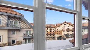 Une vue à travers la fenêtre d’un immeuble doté d’un balcon, avec de la neige au sol et d’autres bâtiments en arrière-plan.