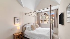 Un lit à baldaquin dans une pièce dotée d’un plafond en pente, une petite table de chevet avec une lampe, une estampe botanique encadrée au mur et une vue sur un balcon avec un bâtiment en arrière-plan.