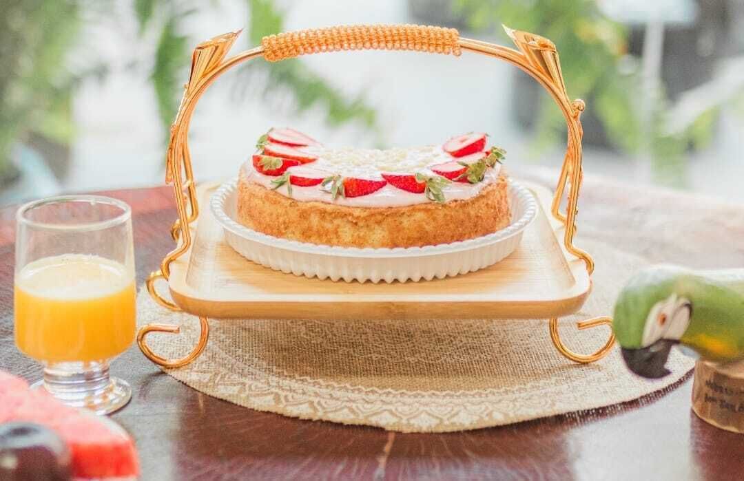 A cake on a wooden stand with a woven basket handle, a glass of orange juice, and a parrot on a table.