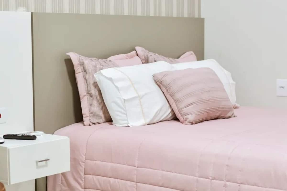 Uma cama com um espesso edredom rosa e almofadas, uma mesa de cabeceira branca com um controle remoto, e uma parede listrada ao fundo.